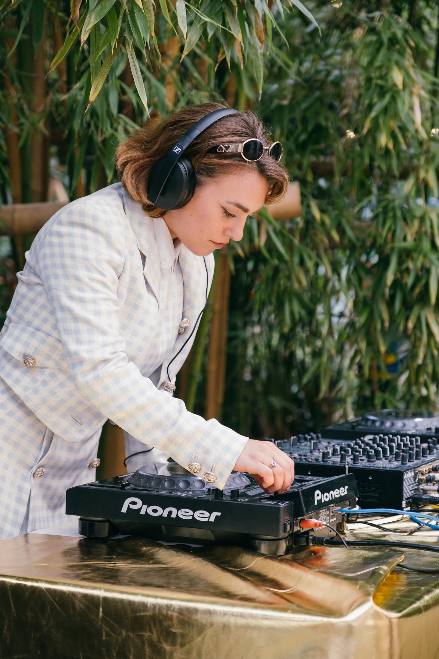 A person wearing a light blue and white checkered suit operates DJ equipment on a gold-colored table outdoors. The individual has headphones on and sunglasses resting on their head while adjusting controls on a Pioneer DJ mixer. The background features tall green bamboo plants, creating a natural setting for the performance.