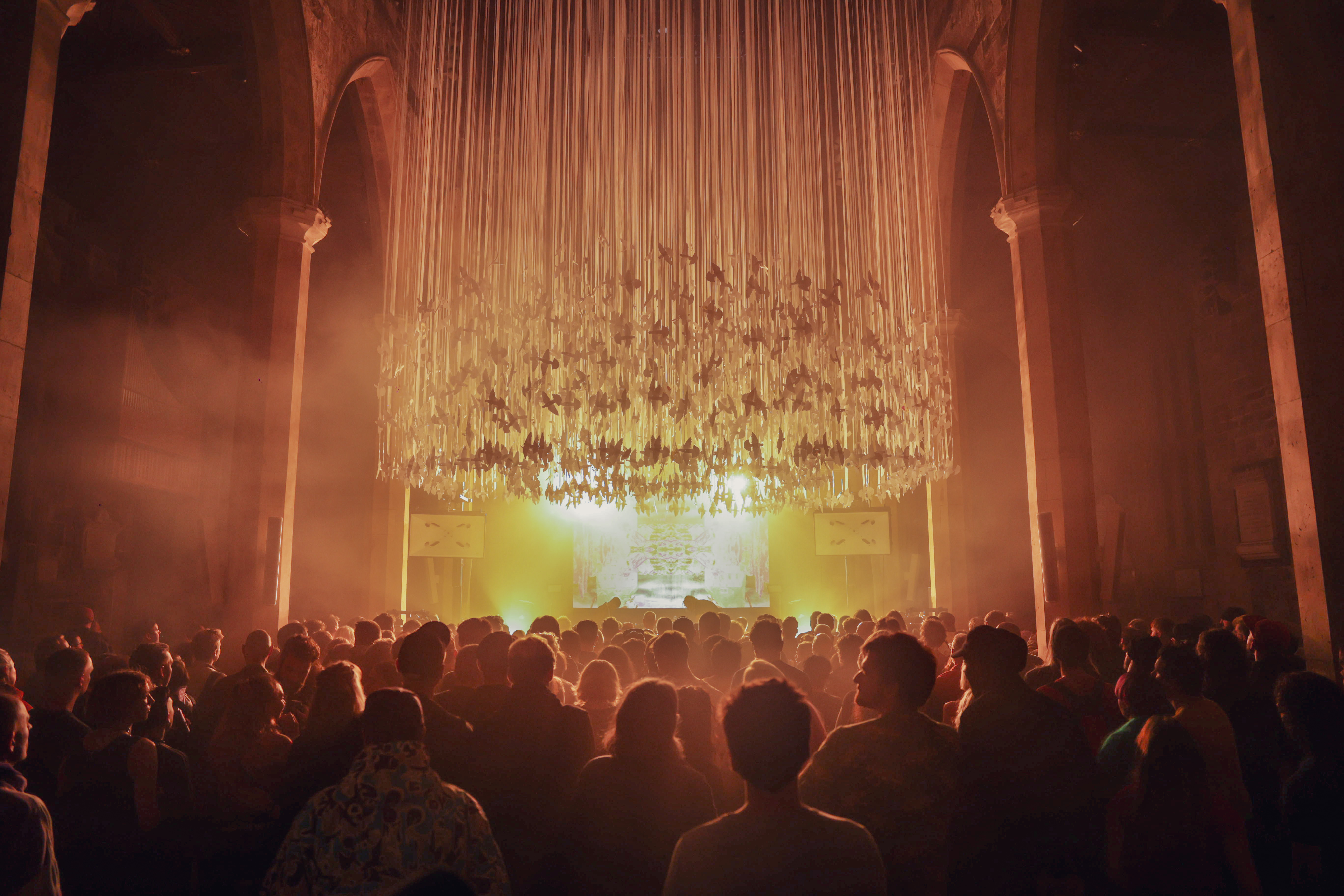 A large crowd gathered inside a grand hall with tall stone arches, facing a brightly lit stage. Above the audience hangs an elaborate installation of long, vertical ribbons or strands, illuminated by warm golden light that fills the space with a dramatic glow. The atmosphere suggests a concert or immersive art event in a historic architectural setting.