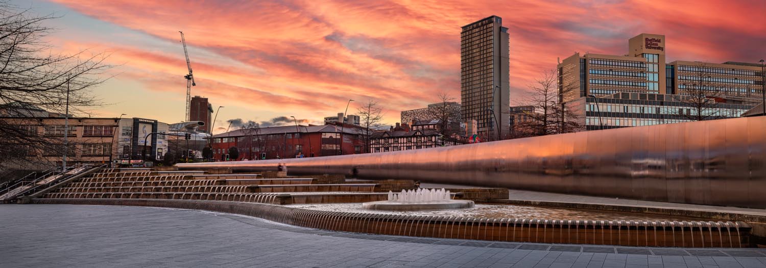 Urban plaza with cascading water features and fountains in front of a long reflective wall. Modern high-rise buildings and older brick structures are visible in the background, along with construction cranes. The sky is filled with dramatic orange and pink clouds during sunset, creating a vibrant backdrop.