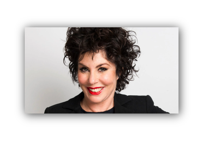  Person with short, dark, curly hair wearing a black blazer over a dark top, posed against a plain light gray background.