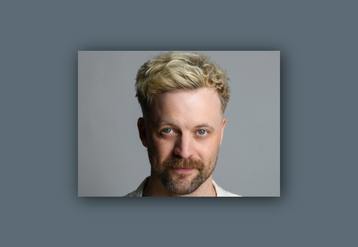 Close-up of a person with short, tousled blonde hair against a plain gray background. The individual is wearing a light-colored shirt.