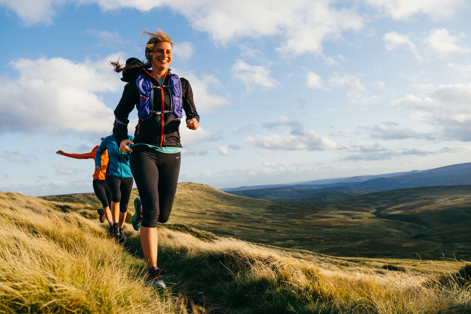Group of people trail running along a grassy ridge in open countryside. The lead runner wears a black jacket, black leggings, and a purple hydration pack. Behind, others in bright athletic clothing follow along the narrow path. Rolling hills and a wide sky with scattered clouds stretch across the background.