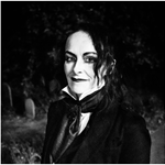 Rosie Garland: A black and white photograph of a person with long, wavy hair, dressed in a dark suit with a high-collared white shirt and cravat. They wear dark eye makeup and lipstick, contributing to a gothic aesthetic. The dimly lit background features gravestones and foliage, enhancing the moody, atmospheric tone of the image.