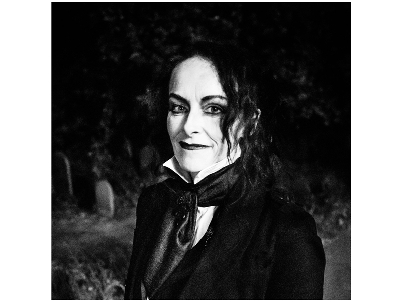 Rosie Garland: A black and white photograph of a person with long, wavy hair, dressed in a dark suit with a high-collared white shirt and cravat. They wear dark eye makeup and lipstick, contributing to a gothic aesthetic. The dimly lit background features gravestones and foliage, enhancing the moody, atmospheric tone of the image.