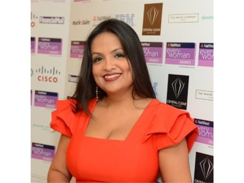 A person wearing a bright red dress with structured, ruffled sleeves is standing in front of a backdrop featuring multiple sponsor logos. The backdrop includes text such as “NatWest everywoman Awards,” “Cisco,” and “Crystal Clear,” along with various brand logos in purple, black, and white. The setting appears to be an event or awards ceremony.