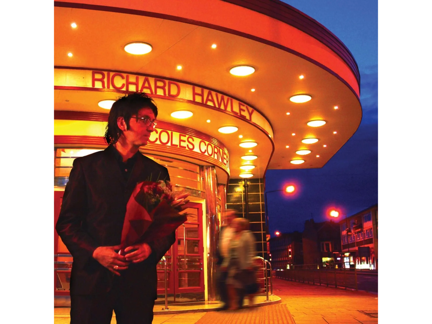 A person dressed in a dark suit stands outside an illuminated art deco-style building at night, holding a bouquet of flowers. The building features a curved canopy with round lights and signage reading “Richard Hawley” and “Coles Corner.” The background shows a deep blue evening sky and blurred figures walking near the entrance.