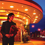 A person dressed in a dark suit stands outside an illuminated art deco-style building at night, holding a bouquet of flowers. The building features a curved canopy with round lights and signage reading “Richard Hawley” and “Coles Corner.” The background shows a deep blue evening sky and blurred figures walking near the entrance.