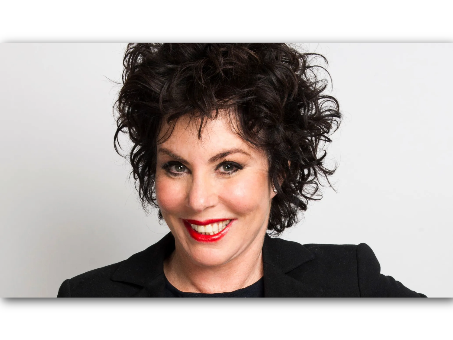  Person with short, dark, curly hair wearing a black blazer over a dark top, posed against a plain light gray background.