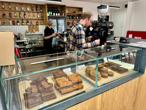 Interior of a café with a wooden counter and glass display case filled with assorted baked goods, including brownies, flapjacks, and cookies. Behind the counter, shelves stocked with bottles and coffee-making equipment are visible, along with a large espresso machine.