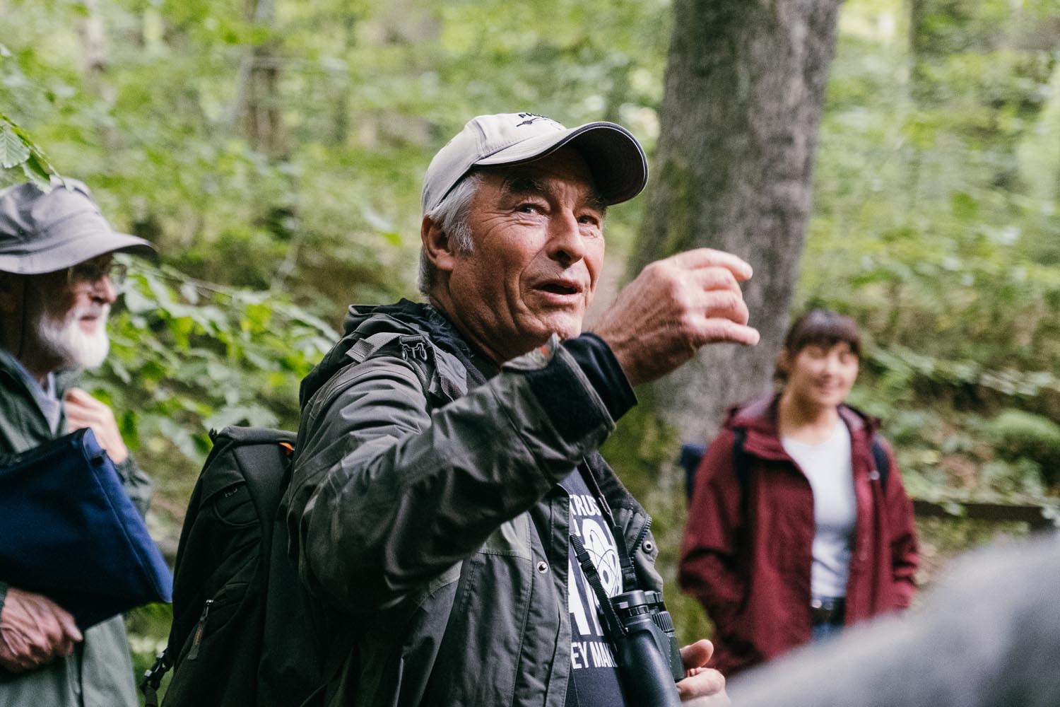 Man talking whilst walking in the woods. A man and woman from the same group can be seen in the background.