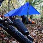 A shelter set up in a wooded area. A bike lies on the ground nearby.