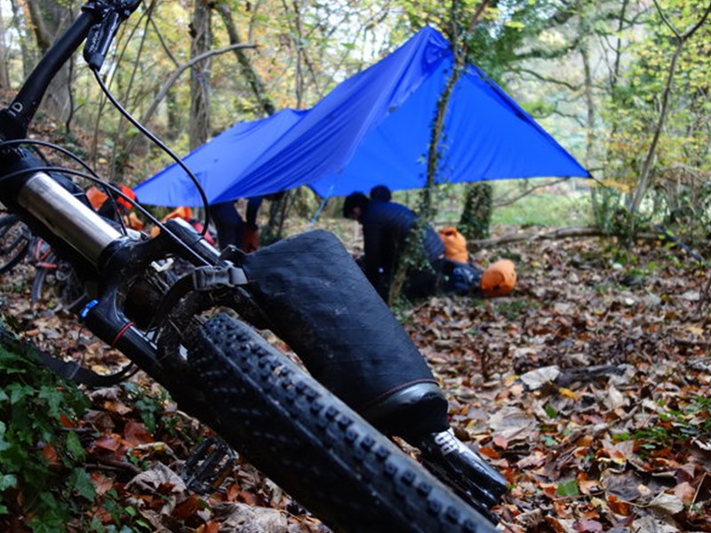 A shelter set up in a wooded area. A bike lies on the ground nearby.