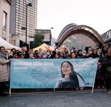 A crowd of adults and children gathered in an urban setting, holding a banner that reads 'Welcome Little Amal, from Sheffield.' The banner includes an illustration of the puppet Little Amal and multilingual messages. Behind the group are buildings and a large arched structure, suggesting a celebratory or welcoming event.