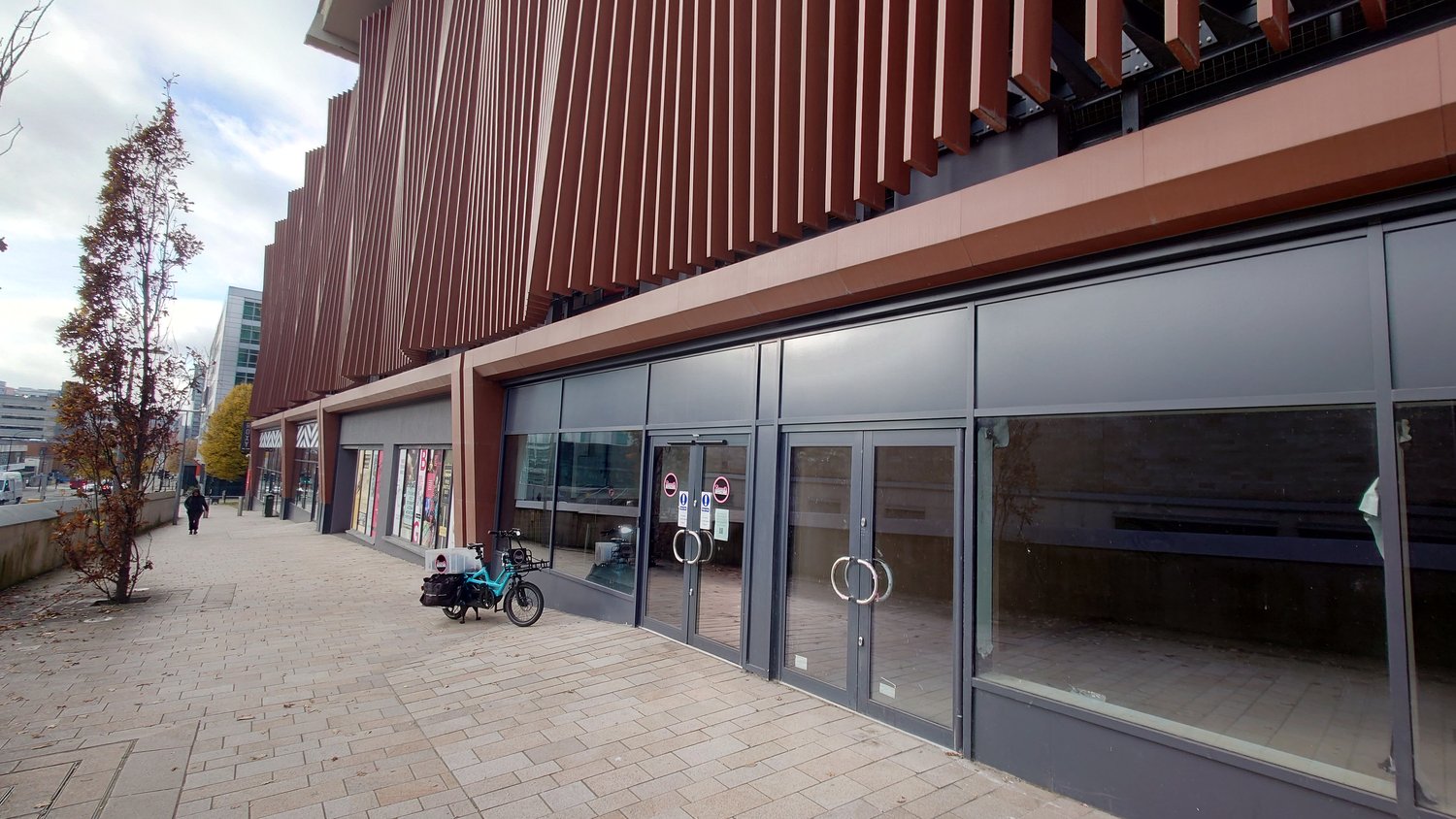 A modern building with tall vertical brown panels and large glass windows along the ground floor. A turquoise cargo bike is parked on the paved walkway in front of the building, and a few pedestrians are visible in the distance. The scene includes a tree with autumn leaves on the left and an urban backdrop with other buildings under a partly cloudy sky.