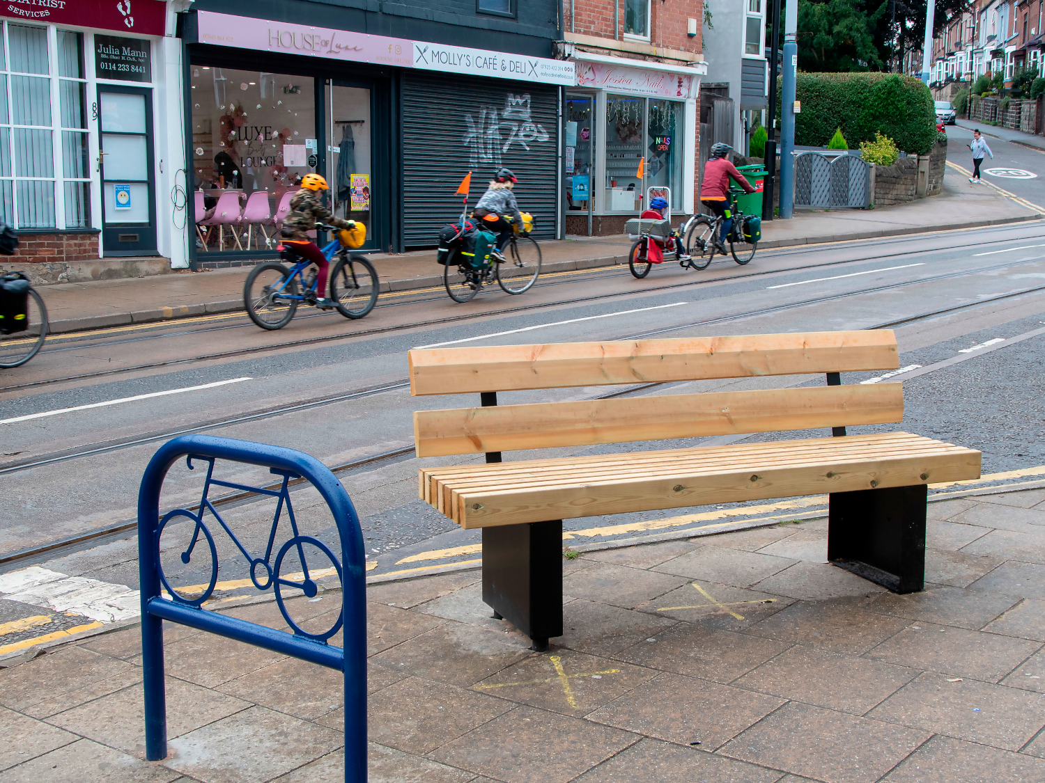 Wooden bench on a paved area next to a blue metal bike rack with a bicycle design. In the background, several cyclists ride along a street with tram tracks, passing shops including “Molly’s Café & Deli” and a beauty salon. The scene shows an urban setting with a slight incline and greenery visible further up the road.
