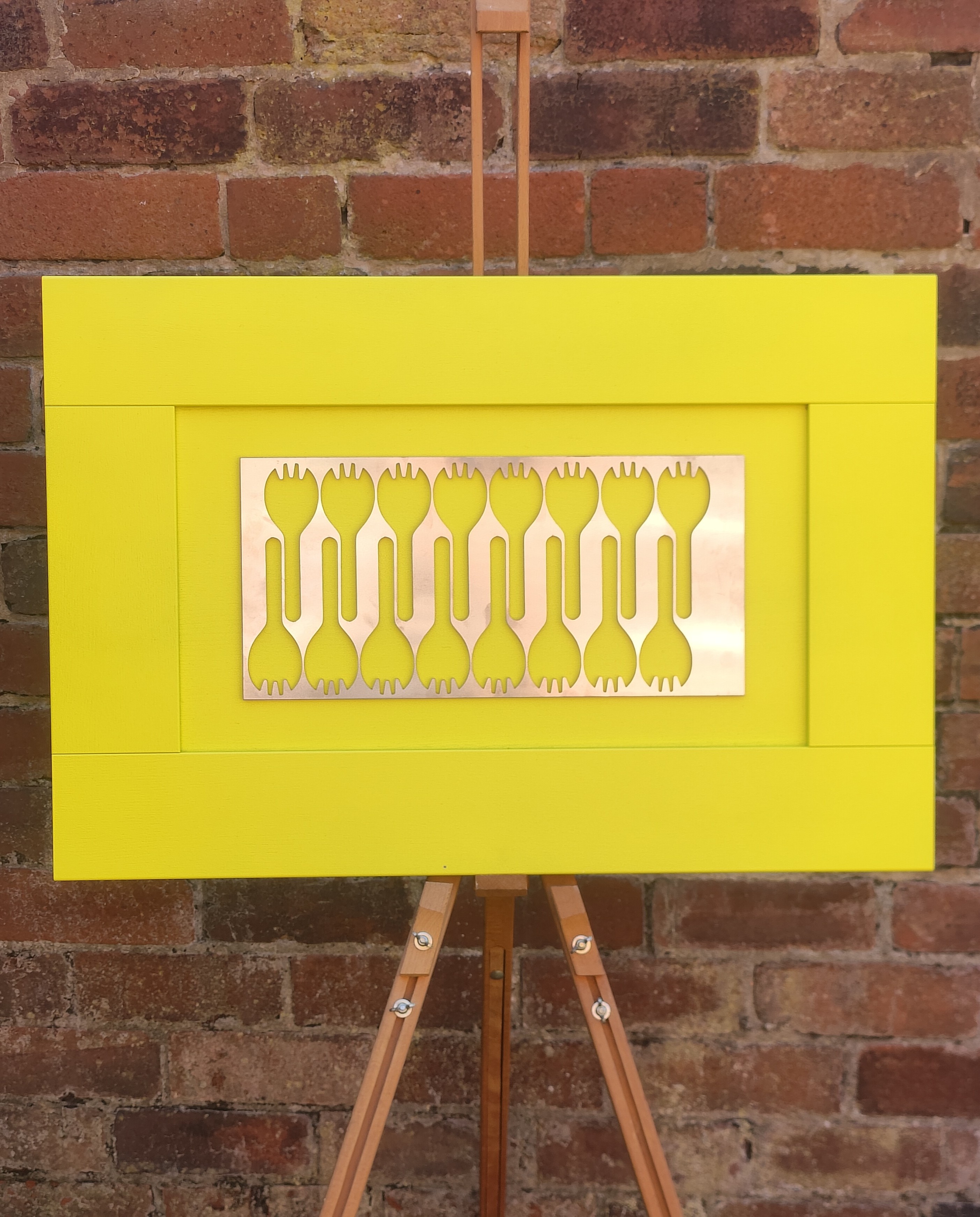Contemporary artwork displayed on an easel, featuring a bright yellow frame and a central metallic panel with cut-out fork-like shapes, set against a brick wall background.