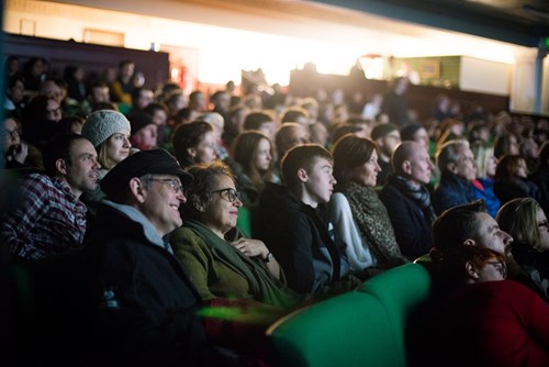 Audience seated in a theater, attentively watching a performance or presentation off-camera. The crowd includes people of various ages in casual attire, with lighting focused toward the stage.
