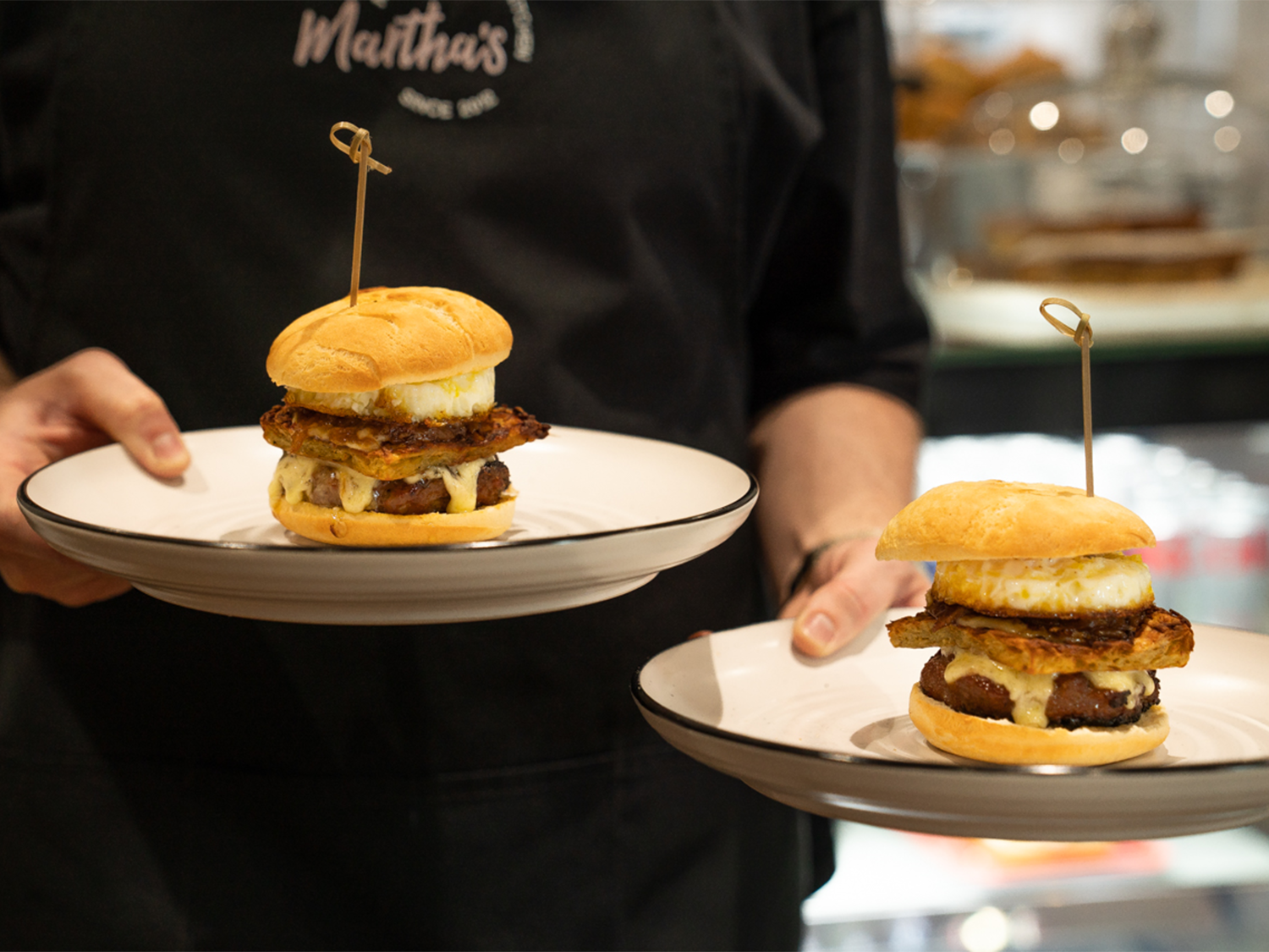 A woman carries two plates, each with a breakfast muffin on.