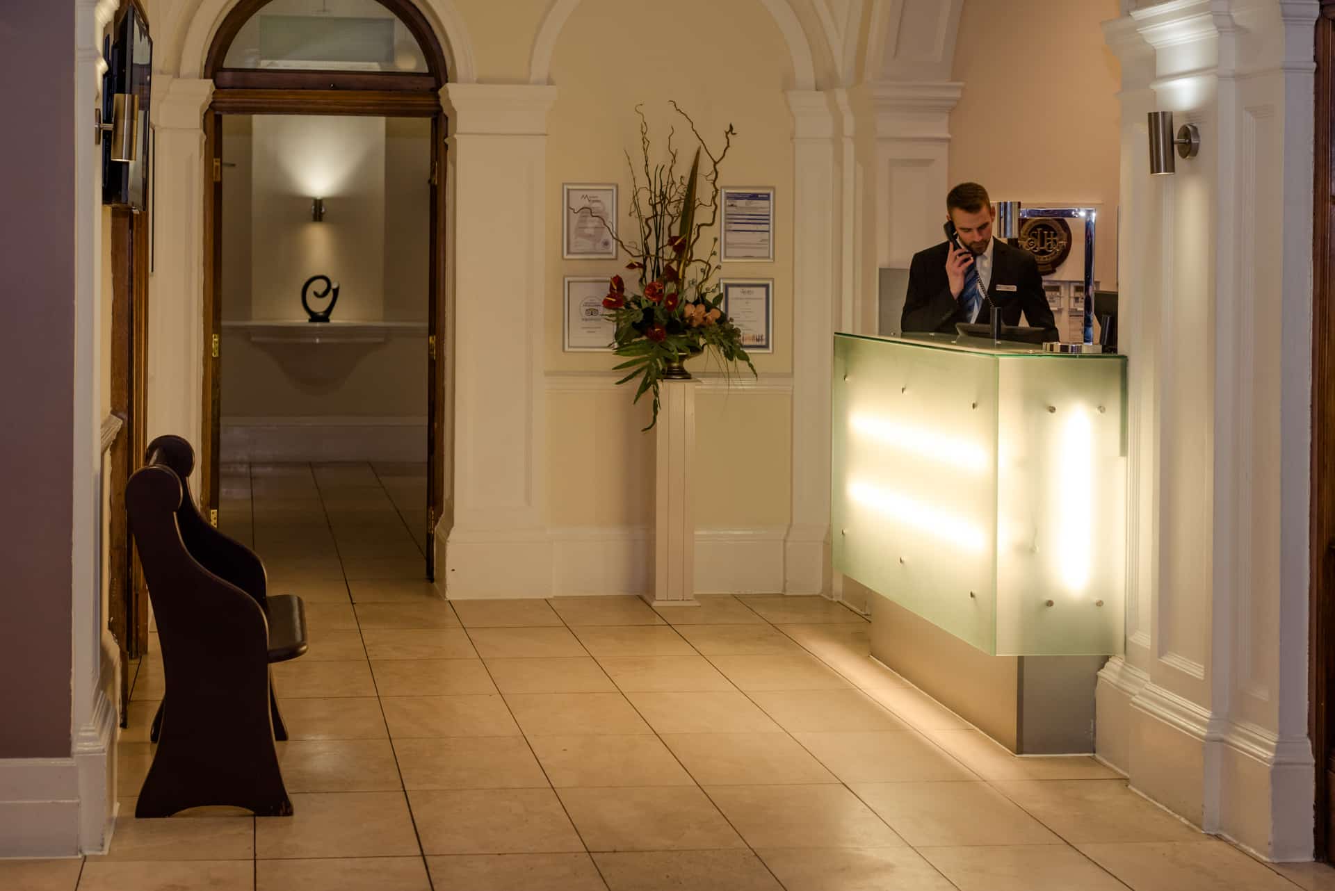 Reception desk in the Leopold Hotel - Reception area of a hotel with a modern glass-front desk illuminated by soft lights. Behind the desk are framed certificates and a tall floral arrangement. The space features cream-colored walls, arched doorways, and tiled flooring. A wooden bench is positioned along the left wall, and an open doorway in the background reveals a decorative sculpture on a shelf.
