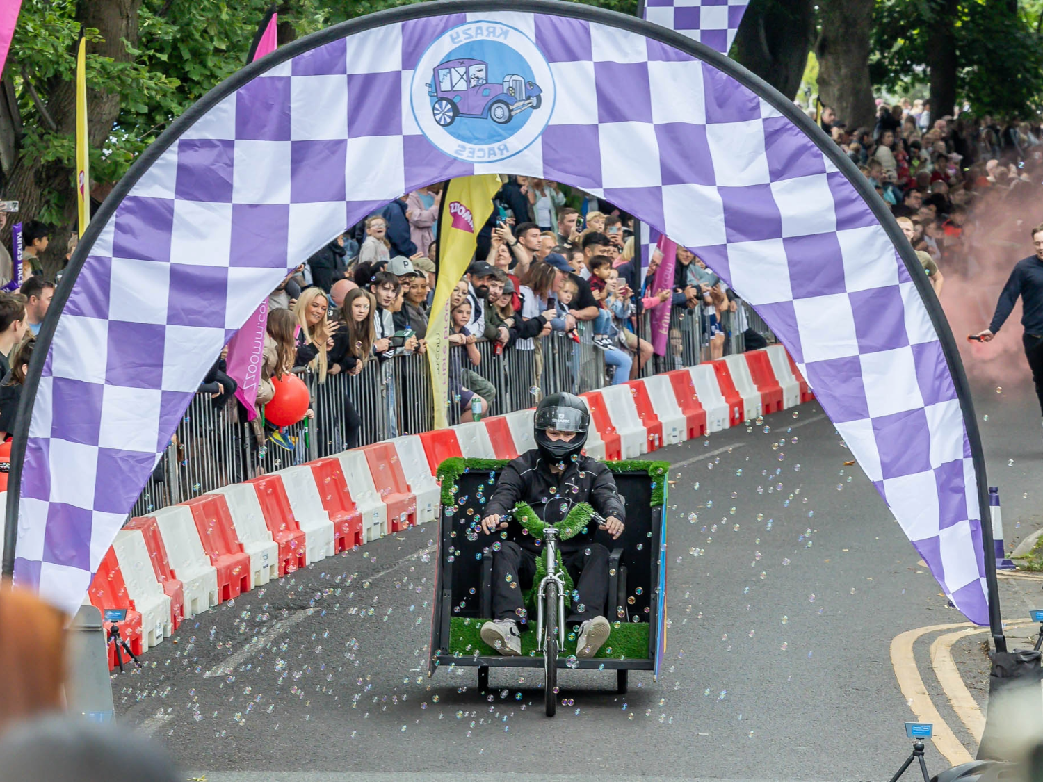 A soapbox racer decorated with artificial grass and blowing bubbles moves downhill under a large purple-and-white checkered archway. The arch displays a circular logo featuring a cartoon car and the words “Krazy Races.” Spectators line both sides of the road behind safety barriers, cheering and holding colourful flags and balloons. Trees and event signage are visible in the background.
