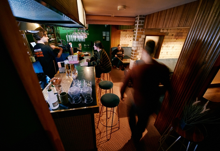 Interior shot of Public, a cocktail bar in the centre of Sheffield. There are people sat in booths and at the bar, where a barman is serving drinks.
