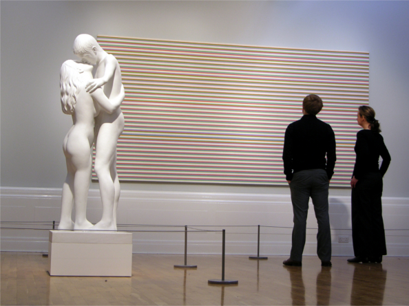 Two people are looking at a painting hung in a gallery. To their left is a marble statue.