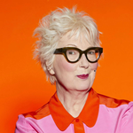 Jenny Eclair is smirking at the camera, she is wearing a pink shirt with a red collar.