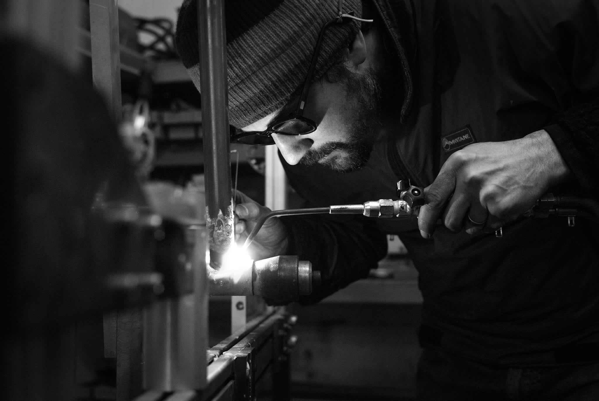 A black and white photo of a man welding a bike frame in a workshop.