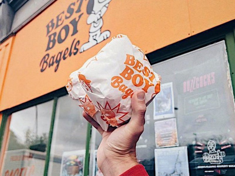 A hand holds a wrapped bagel from Best Boy Bagels, whose sign can be seen in the background.
