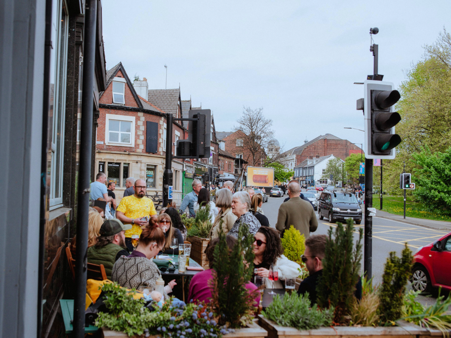 A busy street scene with people gathered at outdoor tables in front of a café or pub. The tables are decorated with planters filled with green shrubs and flowers. In the background, there are traditional brick buildings with shopfronts, a traffic light, and cars on the road. Trees and greenery are visible on the right side under a bright, overcast sky.