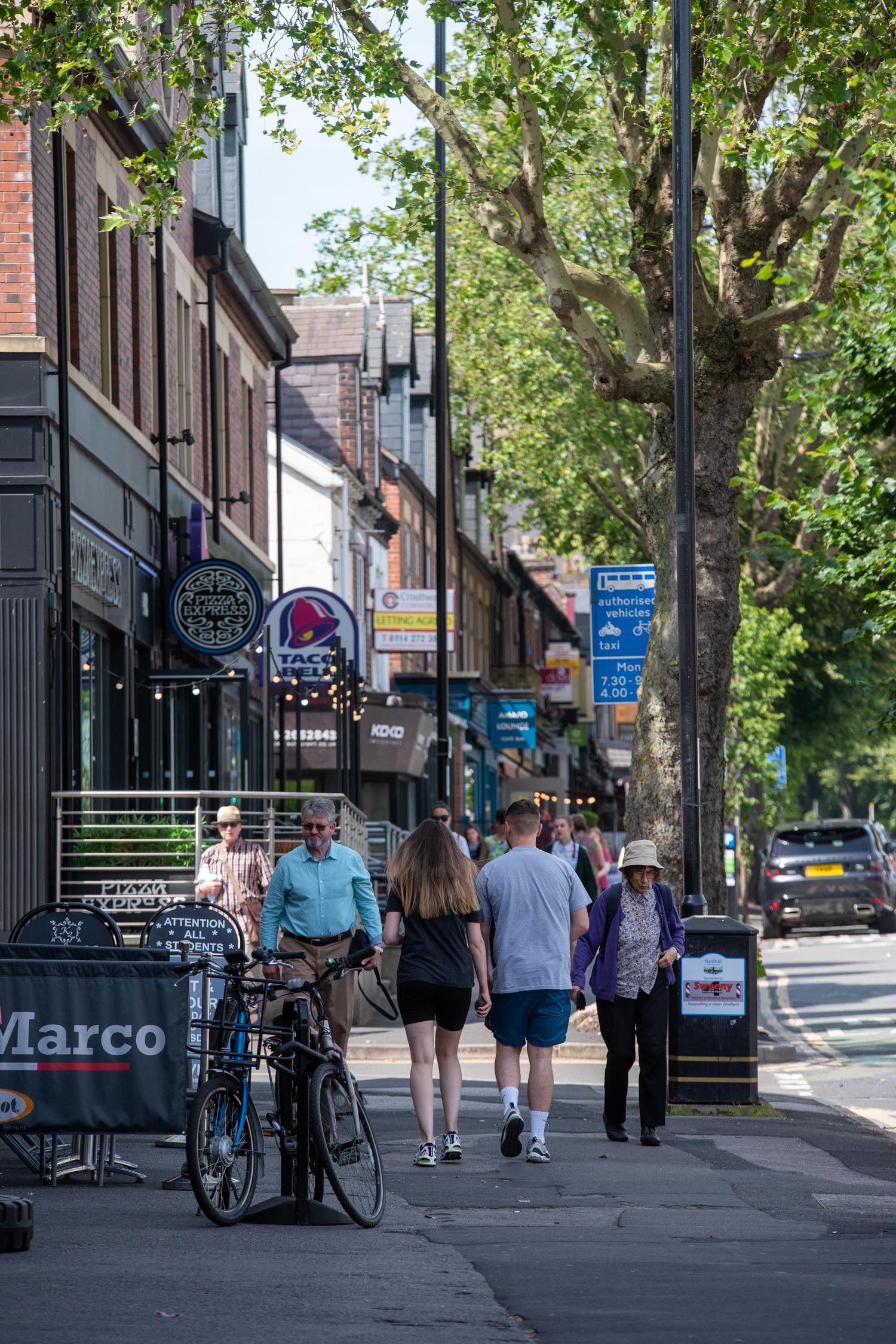 A row restaurants on a tree lined street. There are people walking up and down the pavement.