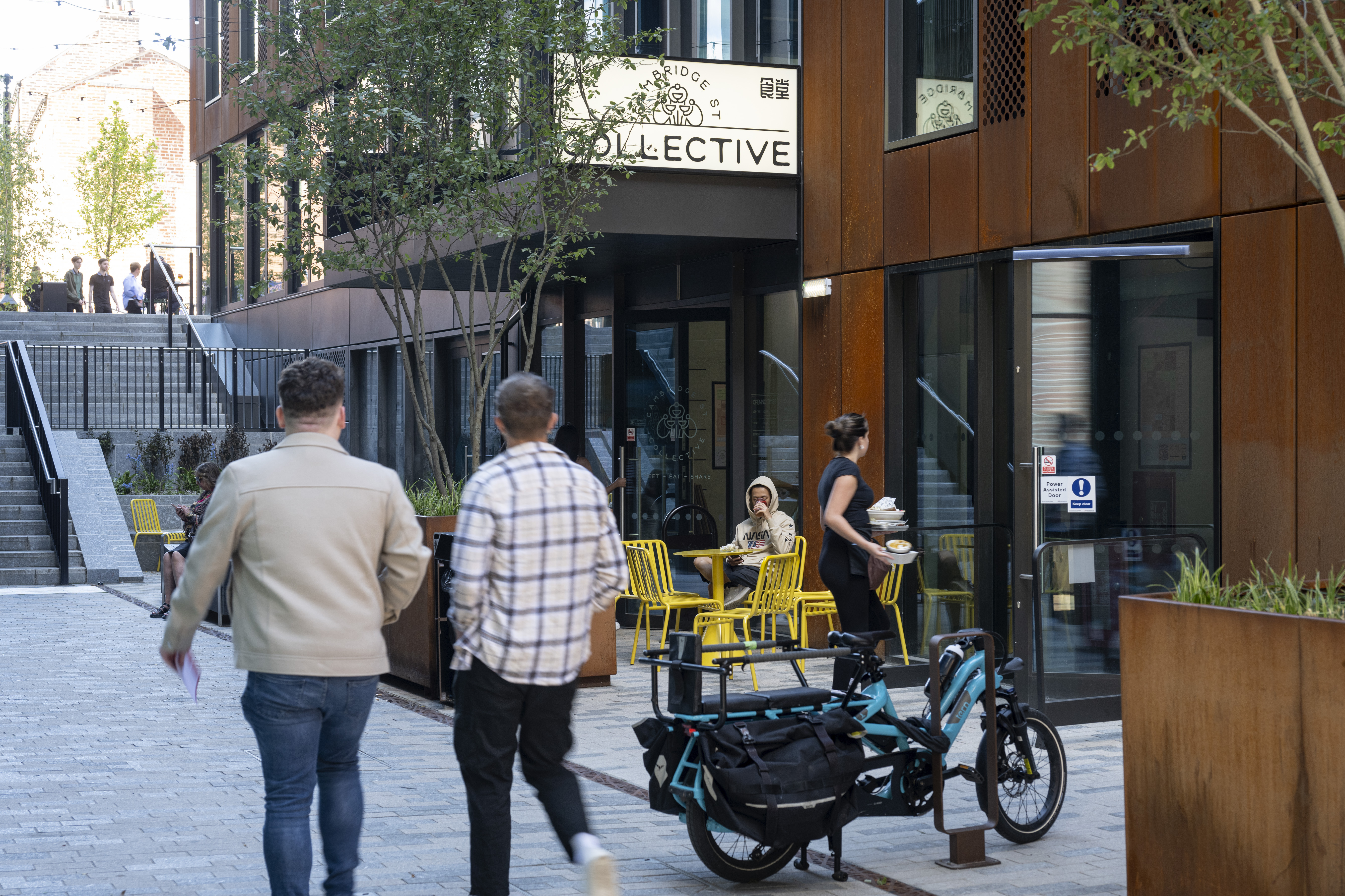 Two people walking along a modern pedestrian street with outdoor seating in front of a café called ‘Collective.’ A cargo bike is parked nearby, and a person is serving drinks to someone seated at a yellow table. The area features contemporary architecture with rust-coloured panels, large windows, and greenery.