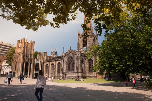 A historic stone cathedral with Gothic architectural features, including pointed arches, tall windows, and a central tower with a clock. The building is surrounded by a paved square with people walking and sitting on the grass. Large trees frame the scene, and sunlight filters through the leaves, casting shadows on the ground. Modern buildings are partially visible in the background.
