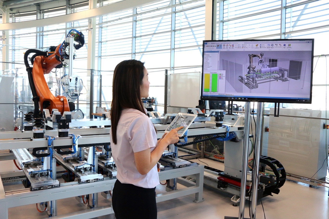 A woman is standing in front of a monitor and holding a tablet. In the background is an industrial robot arm.
