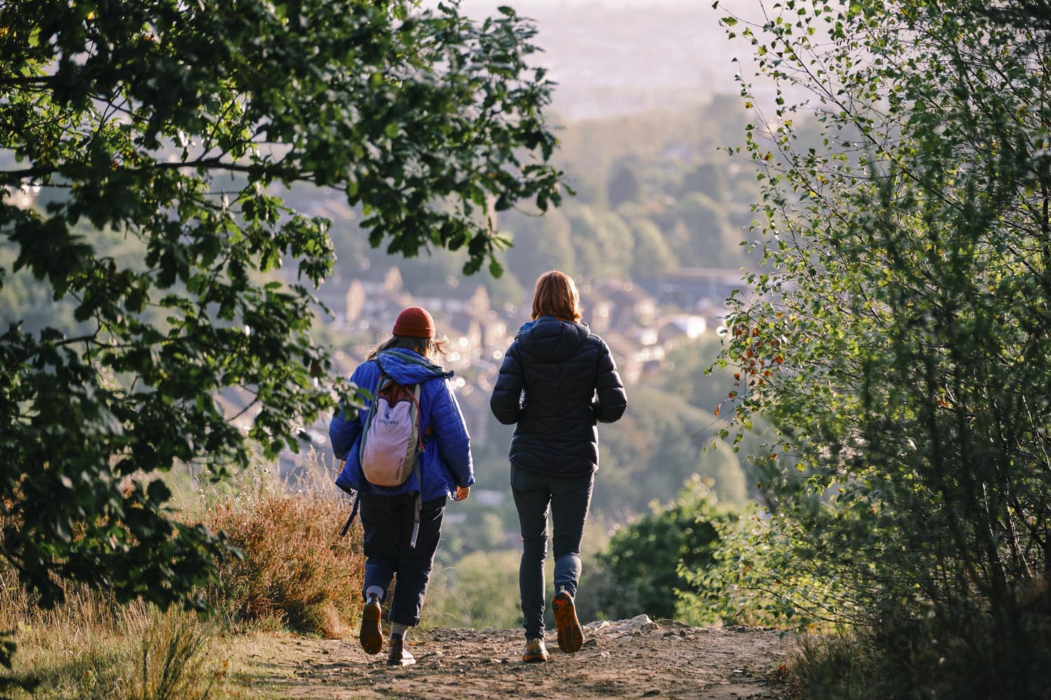 Two people walking along a dirt path surrounded by greenery, with trees framing the scene. They are wearing outdoor jackets and backpacks, and the path leads toward a view of a town nestled among hills in the distance under soft daylight.