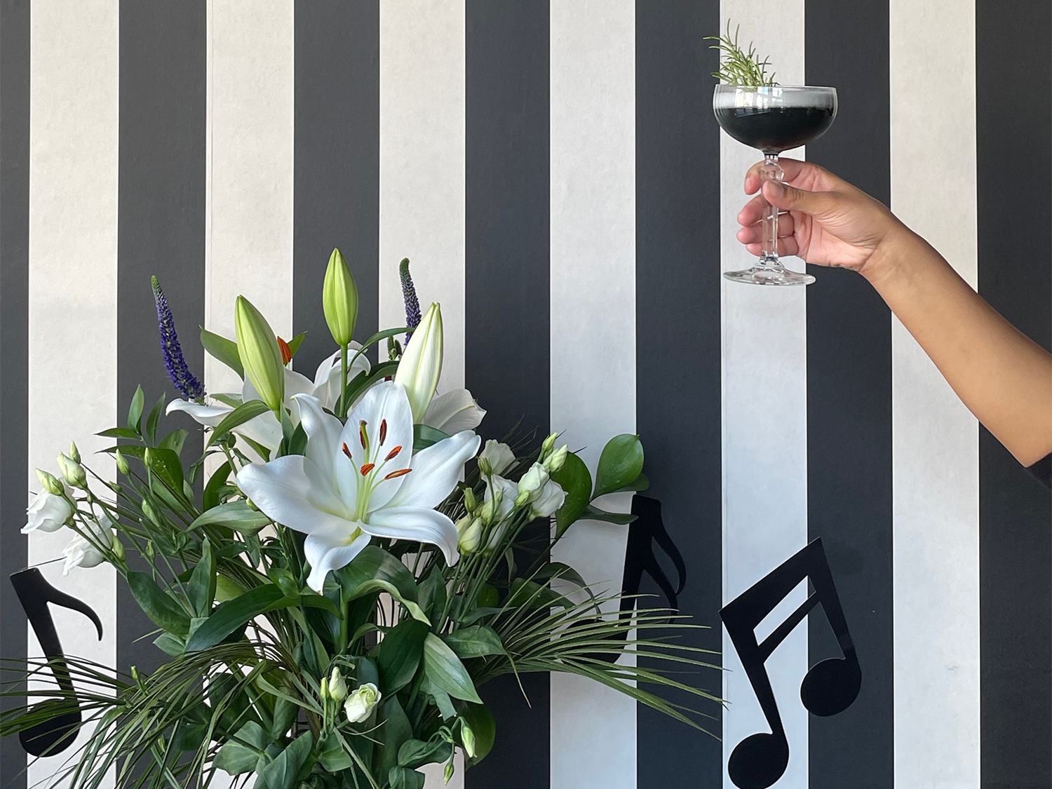 A cocktail held up over a lavish flower arrangement in front of a wall with thick black and white vertical stripes.