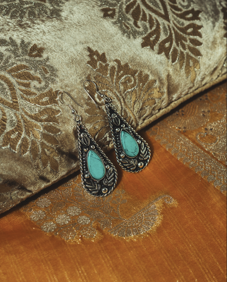 Pair of ornate teardrop earrings with turquoise stones set in intricate silver metalwork, displayed on luxurious gold and bronze patterned fabric.