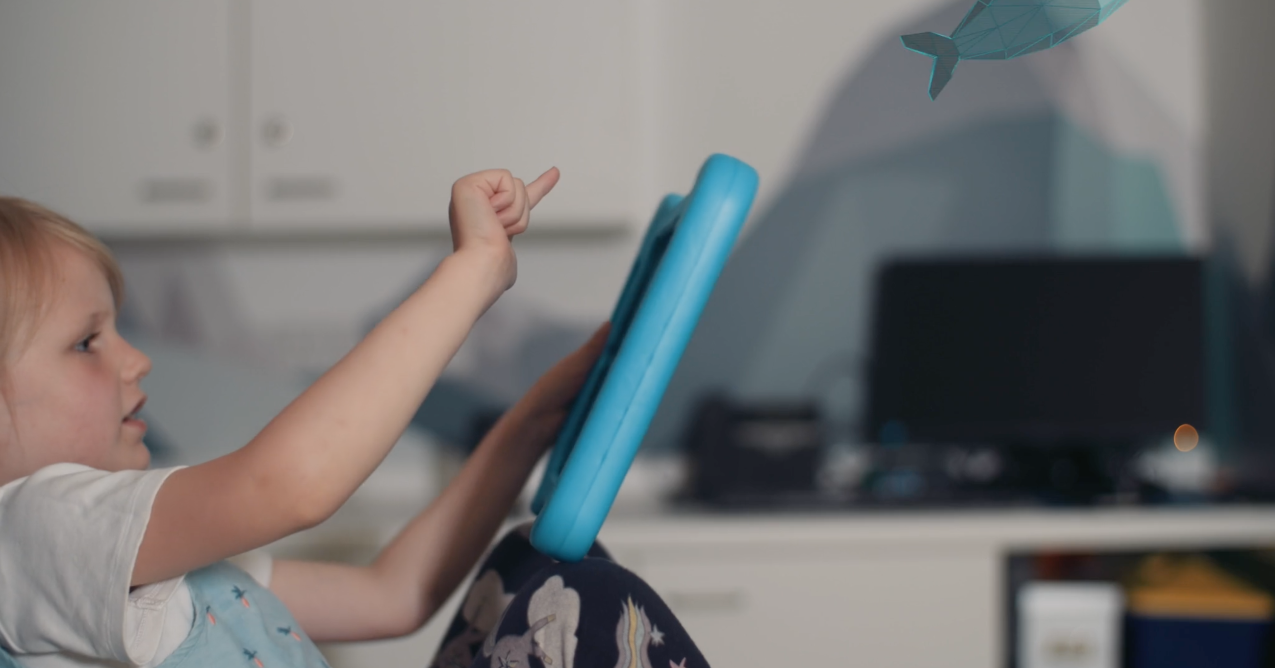 A child is holding a bright blue tablet with one hand while pointing at the screen with the other. The background shows a desk with a computer monitor and office supplies, along with cabinets and a wall featuring geometric designs in muted colours. A paper whale decoration is suspended in the upper right corner of the image.