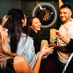 A group of women and men, in their twenties, holding drinks and have a good time while they play darts.