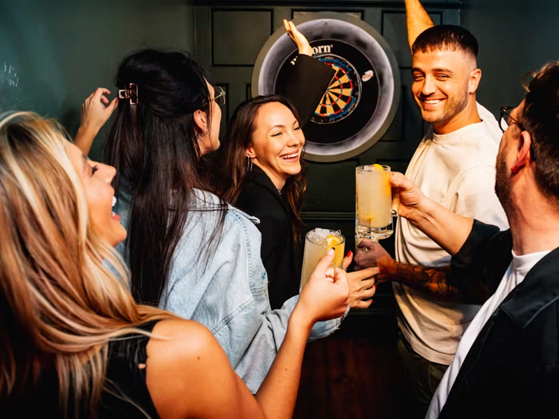 A group of women and men, in their twenties, holding drinks and have a good time while they play darts.