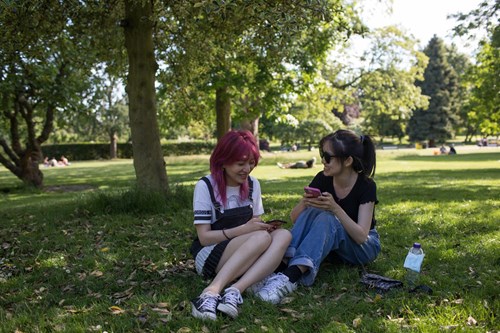 Two people sitting on grass in a park under the shade of trees, looking at their phones. A water bottle and some items are placed nearby, with more people visible relaxing in the background.