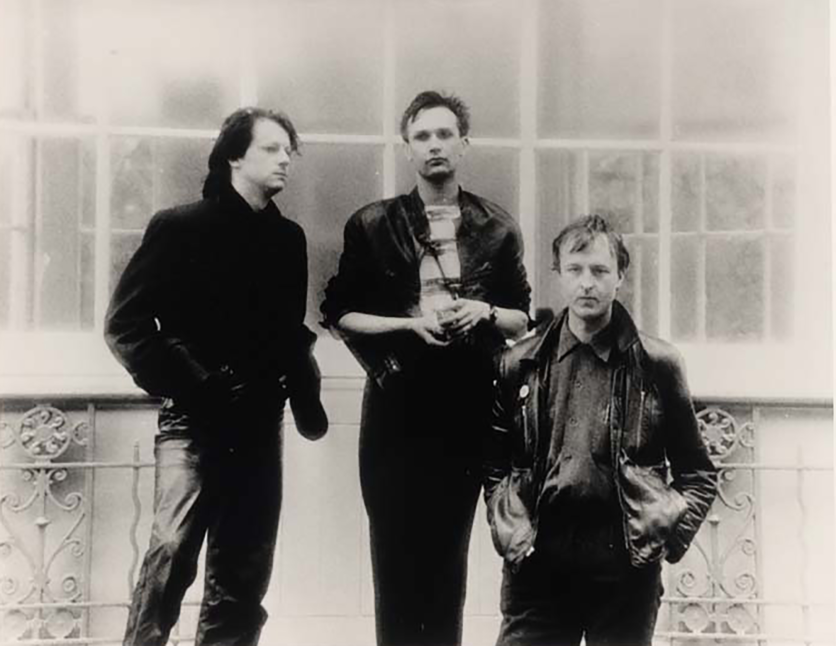 Black-and-white photo of three people standing in front of a large window with decorative railing. They are wearing dark jackets and casual clothing, with hands in pockets or clasped together.