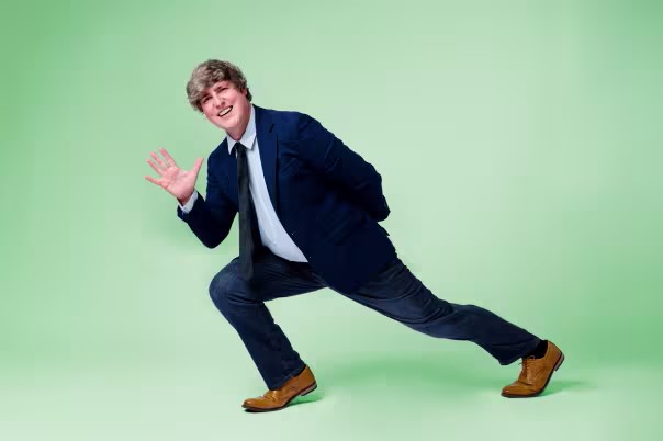 Person wearing a dark blue blazer, white shirt, and dark jeans striking a dynamic pose against a light green background. One leg is bent forward in a lunge, the other extended back, with one arm raised and the other behind the back. Brown dress shoes complete the outfit.