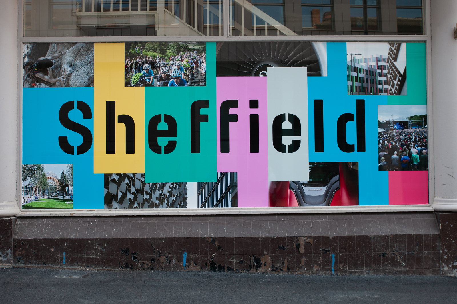 A colorful window display featuring the word “Sheffield” in bold black letters across overlapping panels of blue, yellow, green, pink, and white. Surrounding the text are photographs of city scenes, including architecture, outdoor events, and urban landscapes. The display is set in a large glass window with a dark brown base and a light-coloured frame.