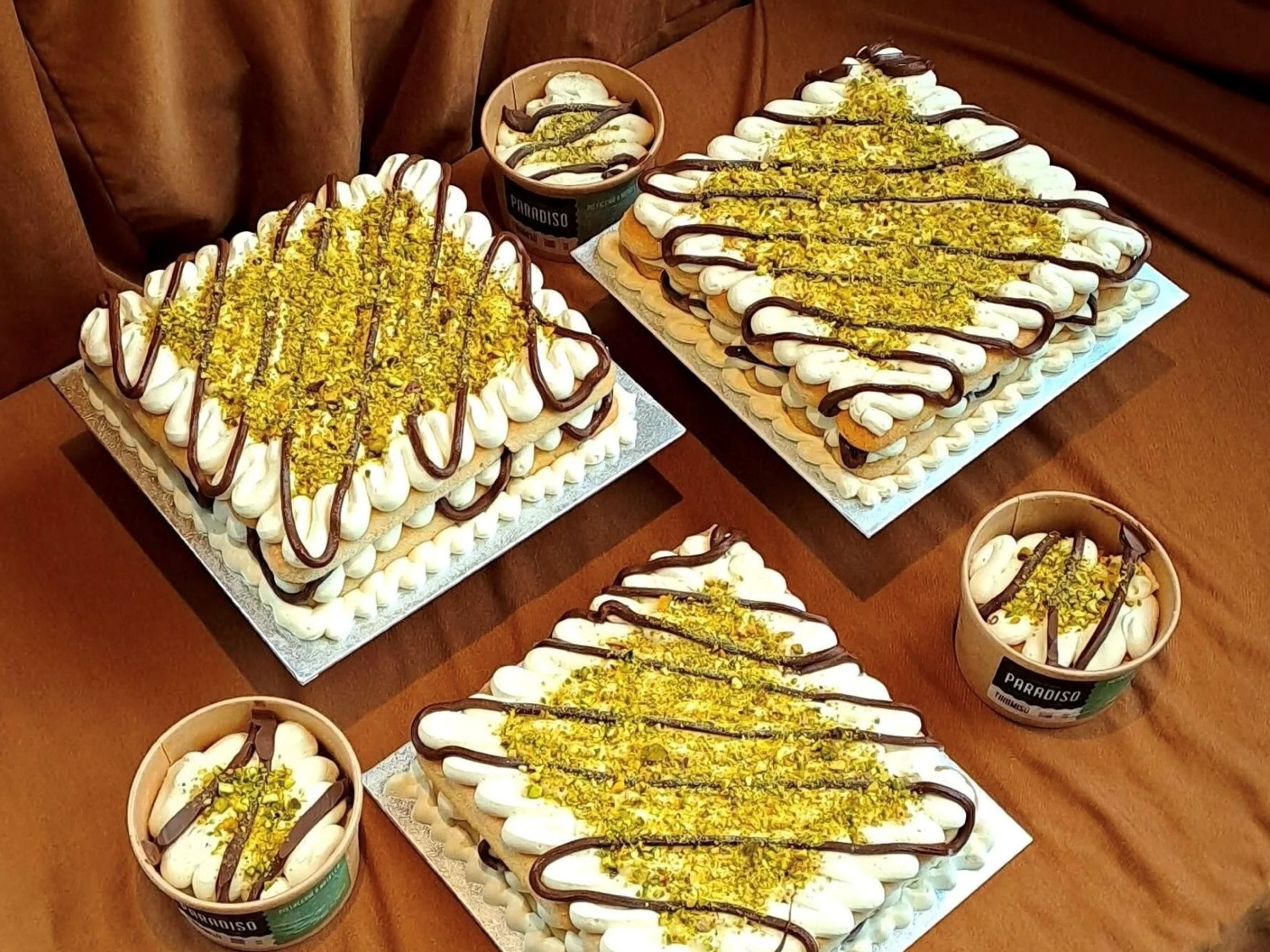 Three square desserts and two round tubs arranged on a brown surface. The square desserts are topped with piped white cream, drizzled with chocolate, and sprinkled generously with crushed pistachios. The two round tubs contain similar toppings with visible cream swirls and chocolate drizzle. All items have a rich, decorative presentation, suggesting indulgent, nutty, and chocolatey flavors.