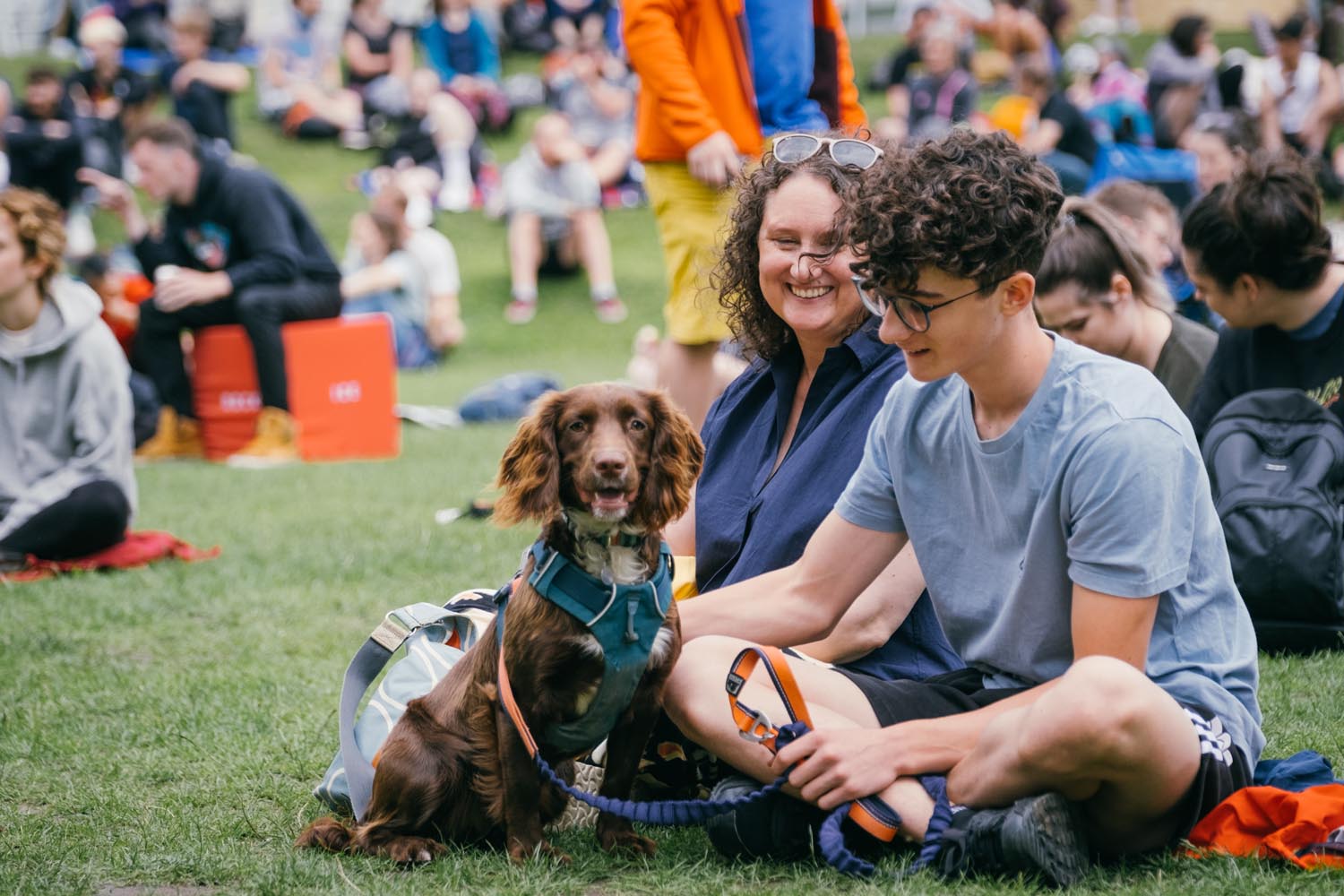 A crowd of people sat on the grass at Devonshire Green at Cliffhanger. In the foreground is a woman, a young man and a dog.