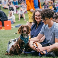A crowd of people sat on the grass at Devonshire Green at Cliffhanger. In the foreground is a woman, a young man and a dog.