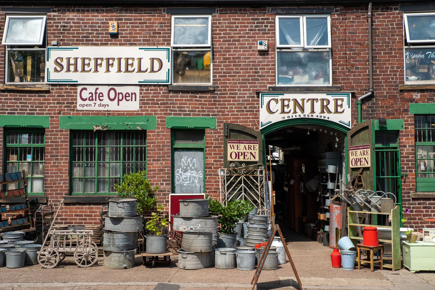 Sheffield Antiques Centre - Brick building with green-trimmed windows and doors, featuring signs that read “Sheffield,” “Cafe Open,” and “Centre.” The entrance has double doors open with “We Are Open” signs. Outside, there are metal buckets, watering cans, and garden tools arranged on the pavement, along with potted plants. The scene suggests a vintage or garden supply shop with a rustic aesthetic.