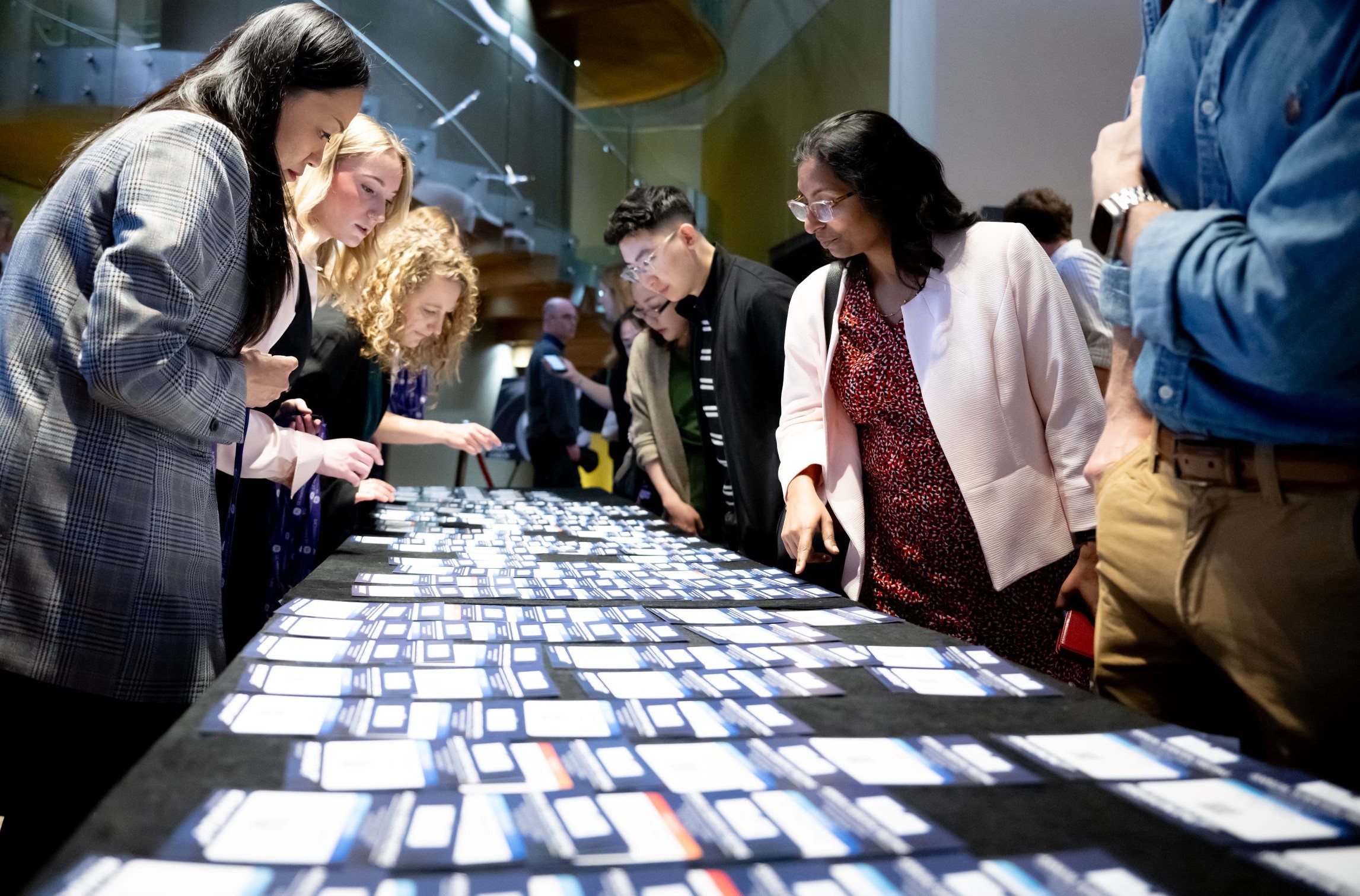 People standing around a long table covered with rows of name badges, selecting or searching for their badges in a modern event registration area.
