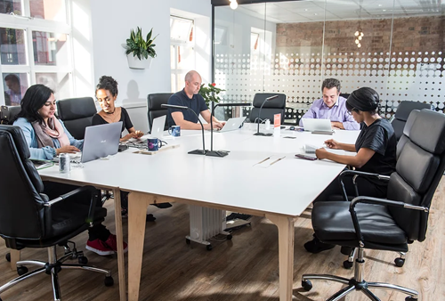 Modern office meeting room with five people seated around a large white rectangular table, each working on laptops. The room features black swivel chairs, wooden flooring, and a glass partition wall with a circular dot pattern. A potted plant is mounted on the wall, and natural light streams in through large windows on the left. The table has a minimalist design with a central stand holding two small decorative items.